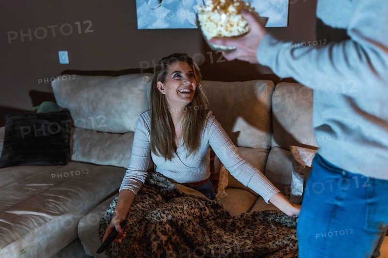 Smiling woman sitting on sofa with remote control, covered with blanket, taking popcorn from boyfriend while watching movie together at home