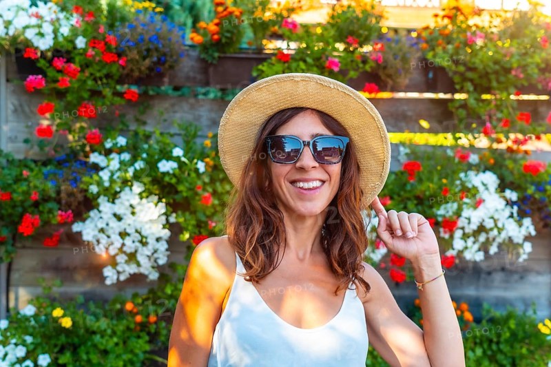 A woman wearing a straw hat and sunglasses is smiling in front of a wall of flowers. The flowers are in various colors and are arranged in a way that creates a beautiful and vibrant display