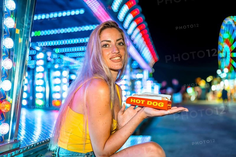 Blonde skinny woman eating a hot dog in an amusement park at night