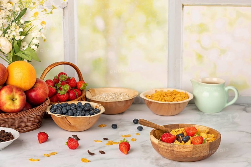 Muesli in a wooden bowl surrounded by ingredients, fruit, strawberries, blueberries, cornflakes, sultanas and oat flakes