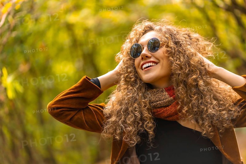 Happy model with curly hair and sunglasses posing and smiling in a park in autumn