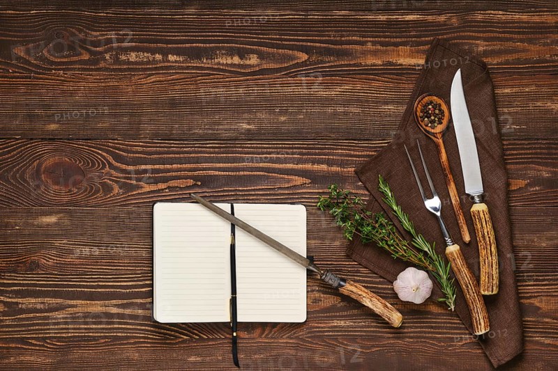 Vintage steak fork, knife and sharpener on wooden table next to notebook. Barbecue appliances and open notepad, food blogging and menu concept