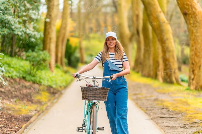 Stylish blonde woman walking with her bicycle on a scenic path, enjoying a leisurely stroll amidst a picturesque park setting