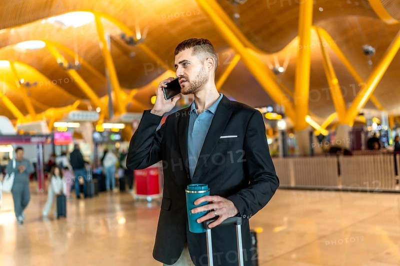 Businessman making a phone call while holding a reusable coffee cup and pulling his luggage at a modern airport terminal