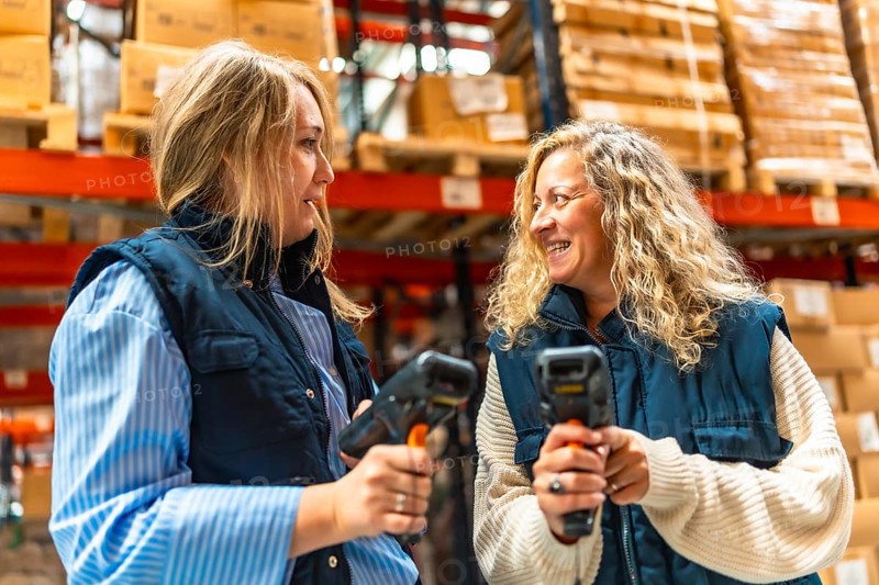 Two female blonde coworkers talking while using barcode scan readers in a distribution warehouse