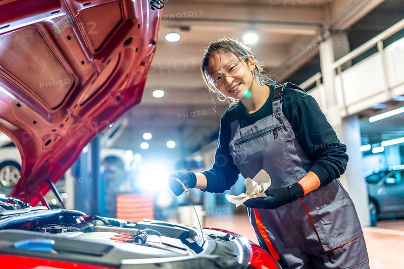 Young female mechanic is checking the oil level of a car engine in a garage, holding a dipstick and a rag