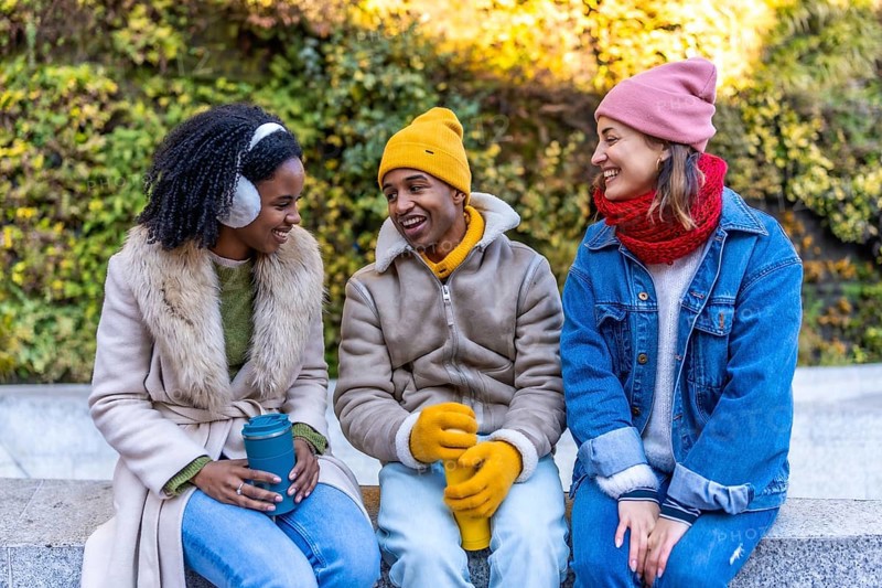 Three cheerful young friends are sitting on a bench in a city park during winter, holding reusable cups and enjoying a conversation