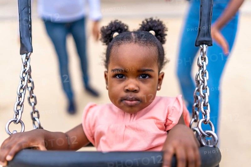 Cute african baby girl sitting on swing in a public park next to her family