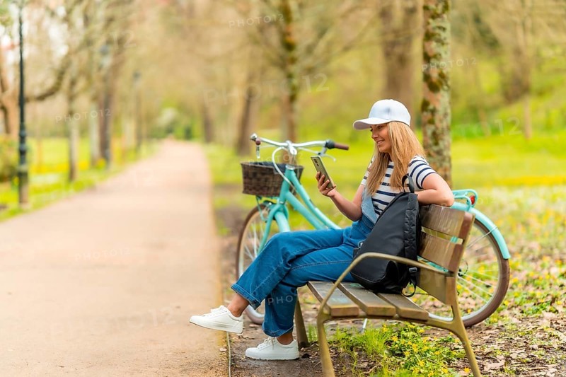 Blonde female tourist wearing casual clothing, using smartphone and resting on park bench with bicycle next to her
