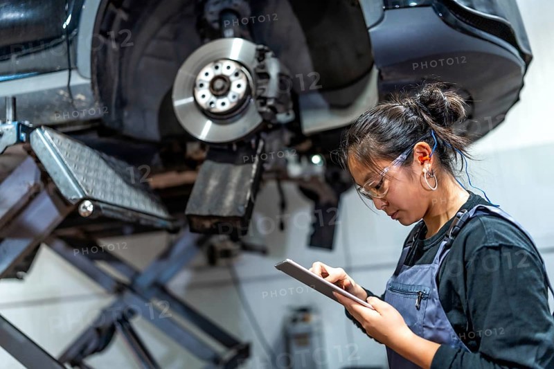 Young female mechanic using a digital tablet while inspecting a car raised on a hydraulic lift in a modern workshop