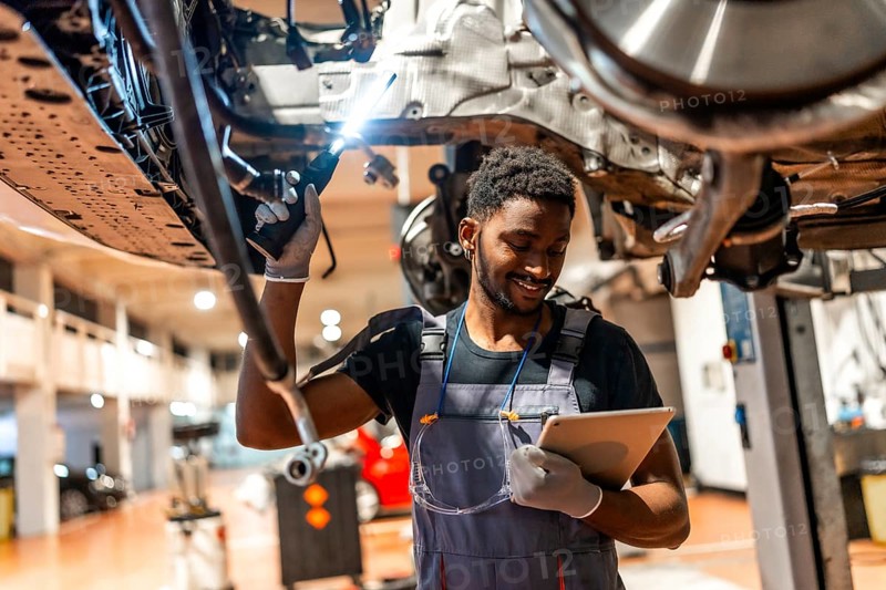 Young mechanic using a digital tablet while working on a car held up by a hydraulic lift in a professional auto repair shop
