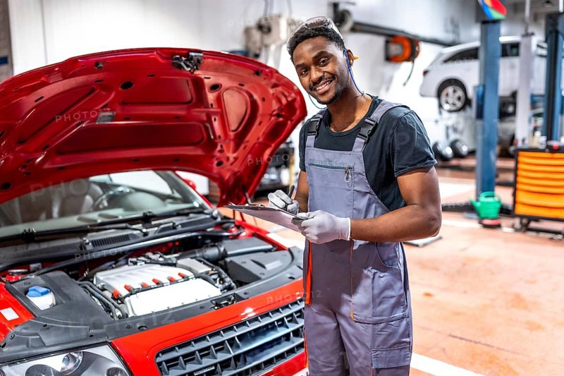 Smiling mechanic taking notes on clipboard while inspecting engine of red car with open hood in a professional workshop