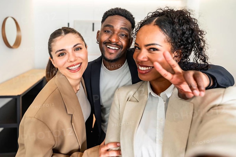 Three cheerful businesspeople taking a selfie, with one making victory sign, celebrating success in modern office