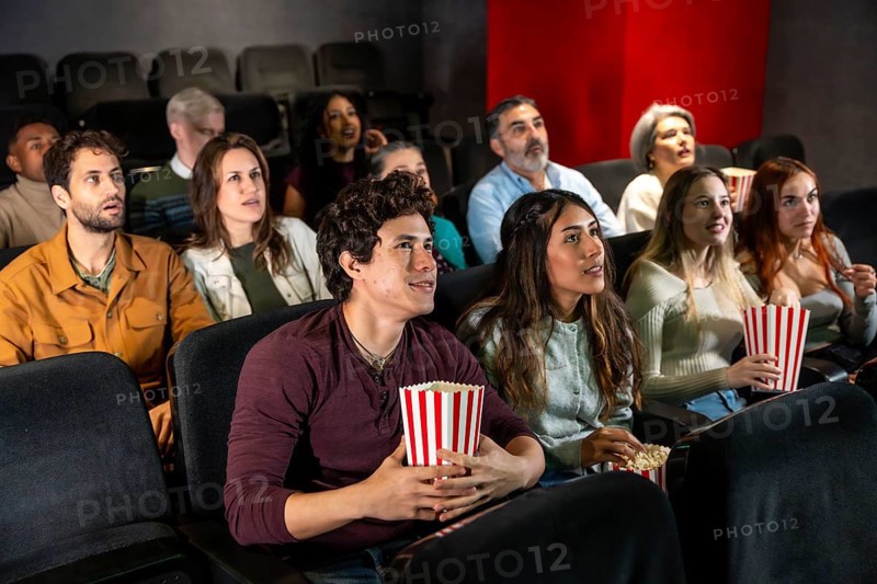 People of different ages and ethnicities are watching a film in a movie theater, holding popcorn buckets and enjoying the show