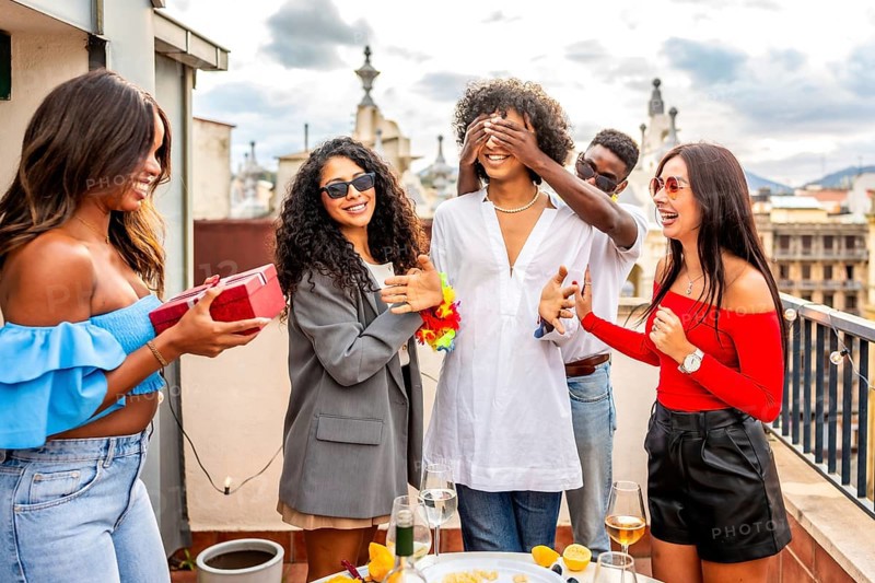 Multiracial group of friends covering eyes of a man giving her a present in a rooftop party