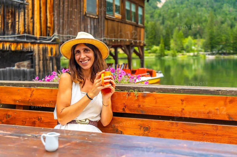 Young woman wearing straw hat smiling and holding a mug in a restaurant by lake dobbiaco with a pedal boat in the background
