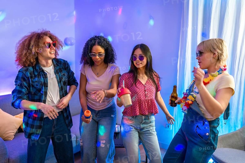 Four friends are singing karaoke and dancing together at a house party, enjoying music, drinks, and colorful lighting