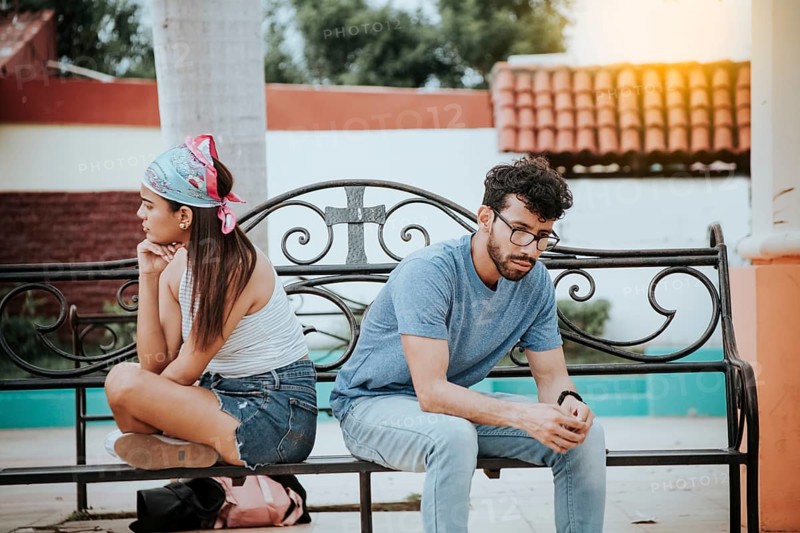 Unhappy couple sitting back to back on bench. Concept of couple problems and crisis. Upset young couple sitting back to back in a park, Young couple arguing sitting back to back on a bench