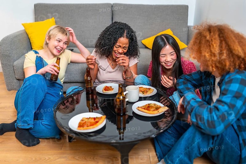 Four female friends sharing a meal of pizza and beer, laughing and having fun together in their living room