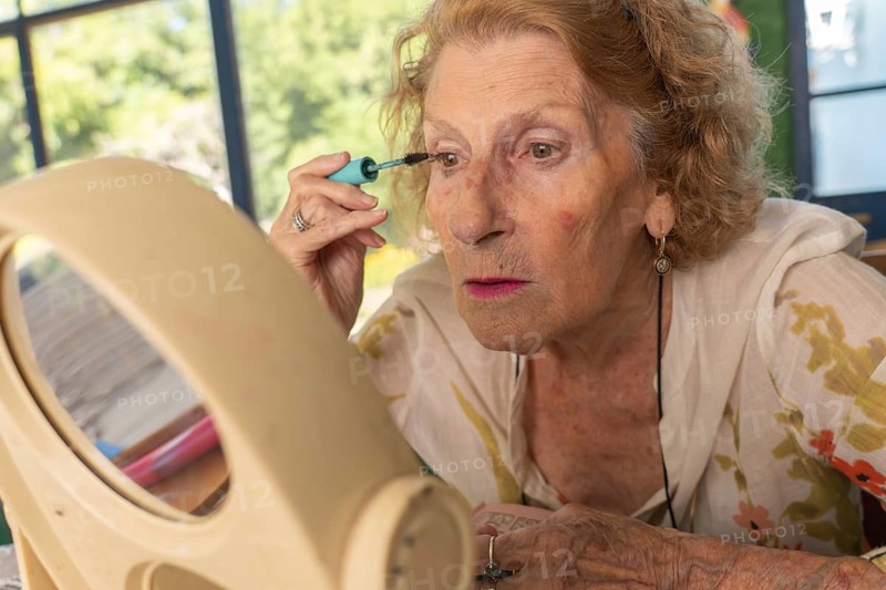 Senior woman applying mascara while gazing into a mirror, preparing for an outing. Embracing her beauty routine, she enjoys a moment of self-care and confidence indoors