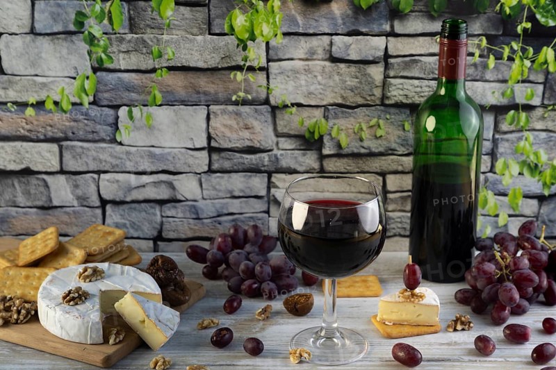 Wine glass and bottle of red wine surrounded by cheese, grapes, nuts and crackers in front of a rustic stone wall