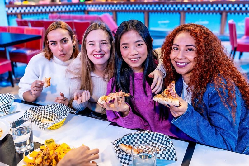 Four diverse young women enjoying a meal together at a restaurant, holding slices of pizza and smiling