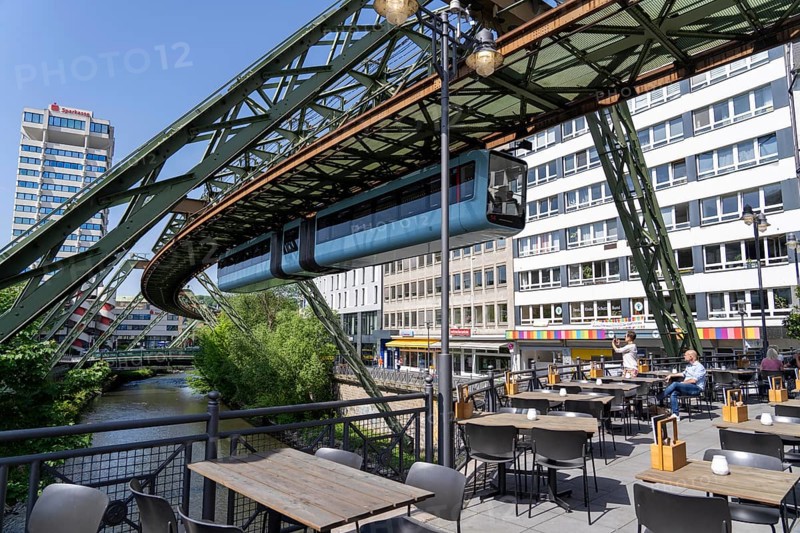 Train of the Wuppertal suspension railway runs along the Wupper, at the main station stop, terrace of a restaurant on a bridge over the river, North Rhine-Westphalia, Germany, Europe