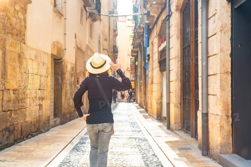 Female tourist wearing a straw hat walking down a narrow cobblestone street in the historic city center of tarragona, spain, enjoying a sunny day of sightseeing