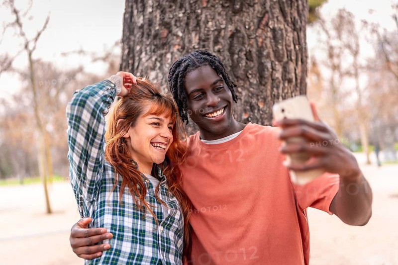 Young multiracial couple embracing and taking a selfie with a smartphone in a park, enjoying a beautiful day together