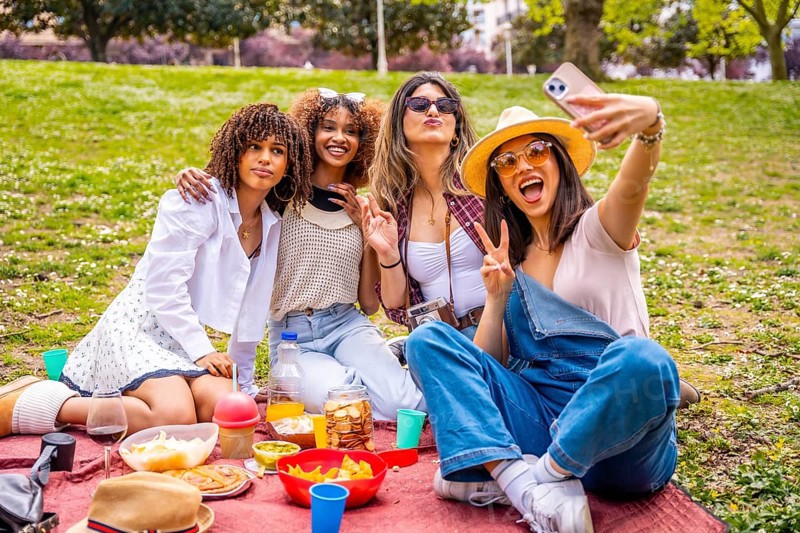 Four cheerful young women enjoying a sunny picnic in the park, smiling and taking a selfie together with a smartphone, capturing joyful moments