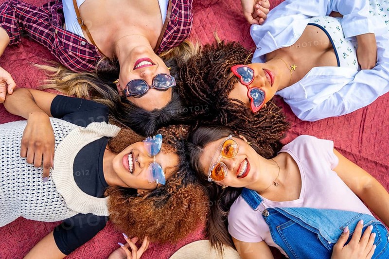 Four diverse young women lying together on a vibrant red blanket, smiling brightly at the camera while enjoying a sunny day outdoors