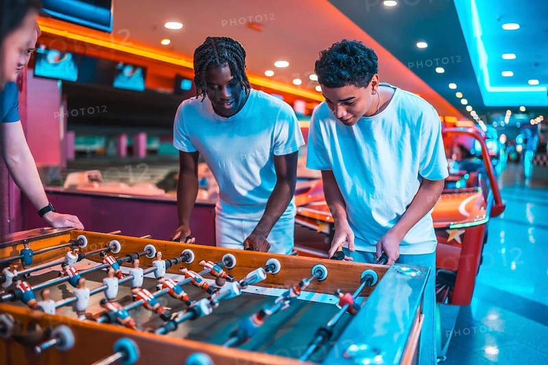Two young men concentrating on playing foosball in a brightly lit entertainment center, enjoying a friendly competition