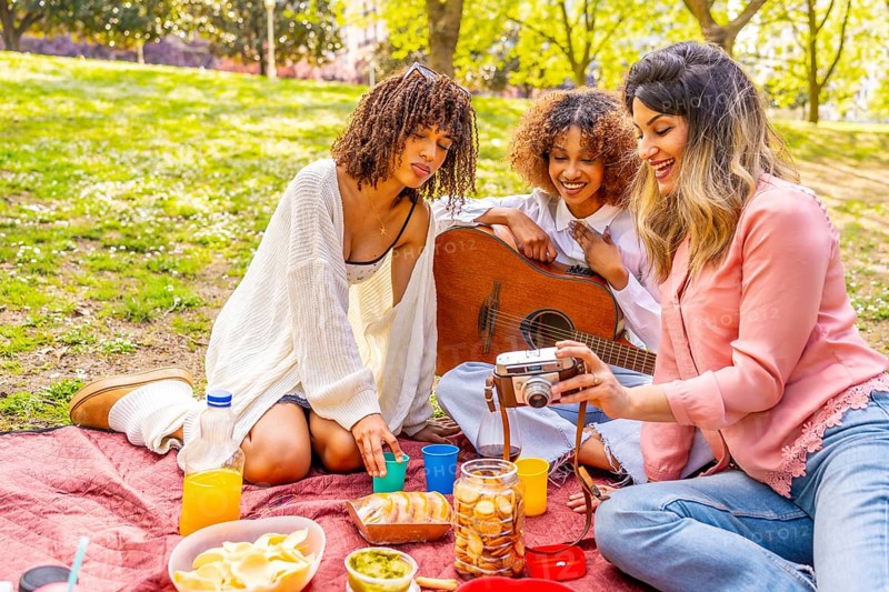 Three female friends enjoying a picnic in a park, taking pictures with a vintage camera, eating snacks, and playing guitar