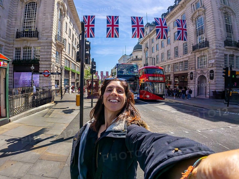 Young woman taking a selfie in london with union jack flags and a red double decker bus in the background, enjoying a sunny day