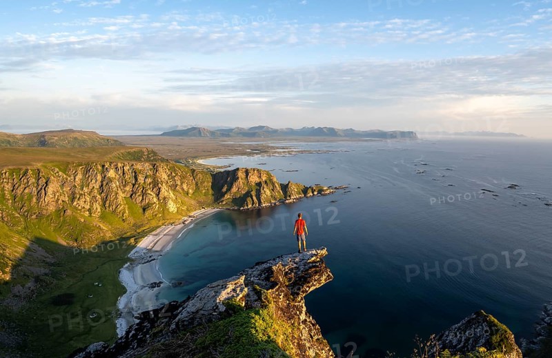 Evening mood, mountaineer standing on a rocky outcrop on the summit of Måtind, view of coastal landscape, Høyvika beach and sea, Måtinden, Andøya island, Vesterålen, Nordland Norway