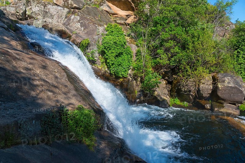 A small waterfall flows over large rocks, surrounded by green plants under a sunny sky, Cascada del Diablo, Devil's Waterfall, Garganta de Gualtaminos, Gualtaminos Gorge, La Vera, Cáceres, Caceres, Extremadura, Spain, Europe