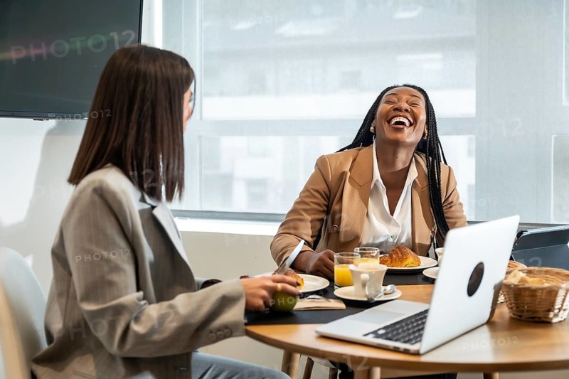 Two businesswomen are enjoying breakfast and laughing together during their business trip, creating a positive and productive atmosphere