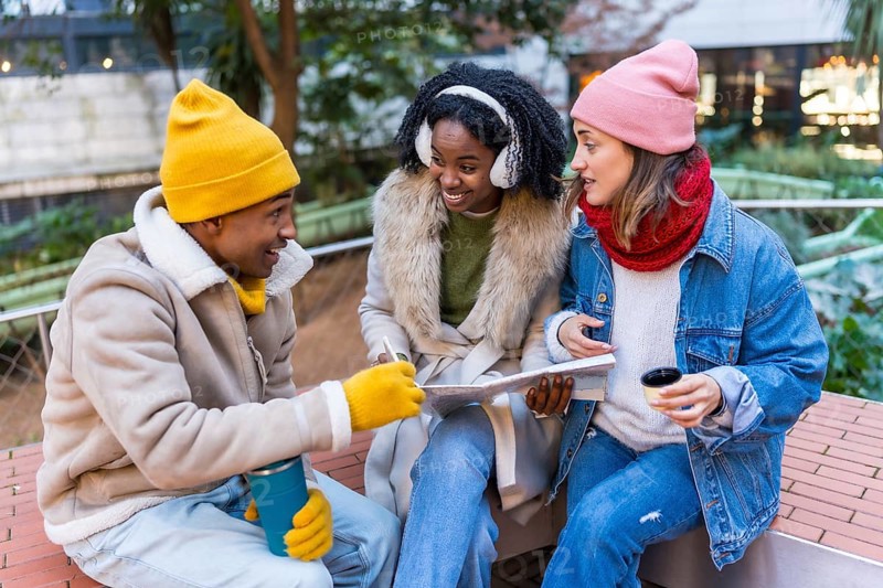 Three happy friends sitting outside in winter, drinking coffee and looking at a map, planning a trip together