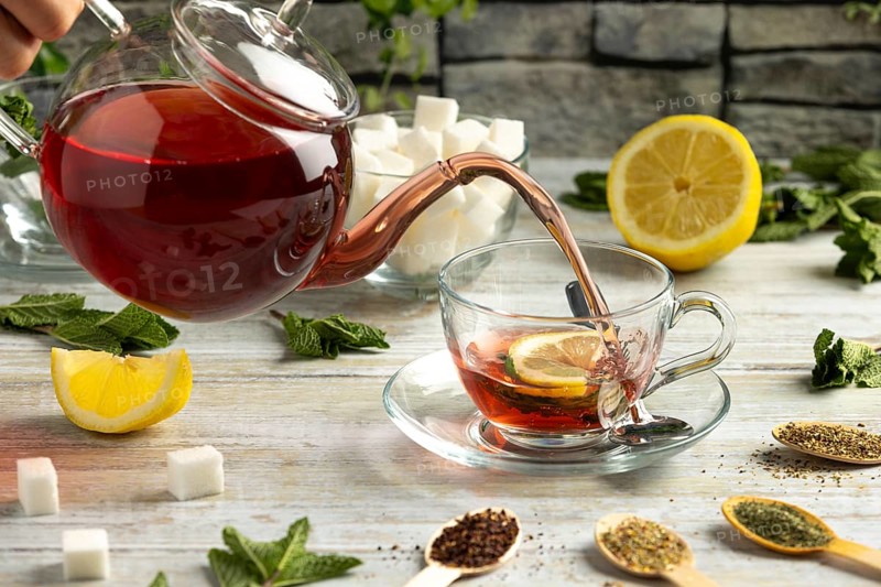 Red tea is poured from a pot into a glass cup with lemon and mint, surrounded by sugar and teas