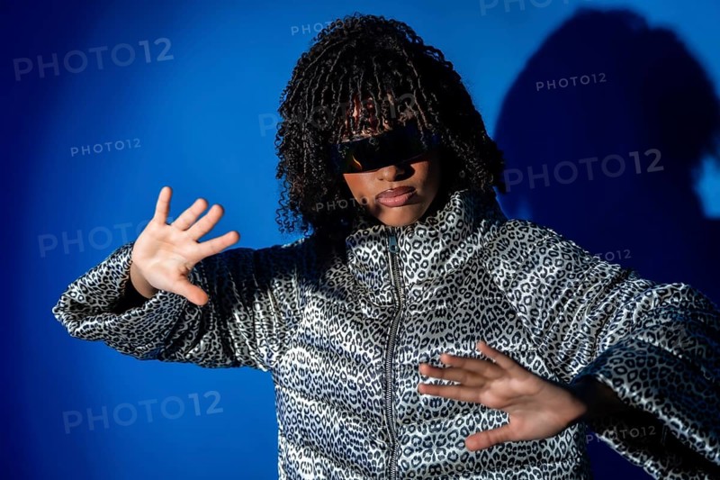 Young woman with curly hair and sunglasses dancing with hands up, wearing a patterned jacket on a vibrant blue backdrop