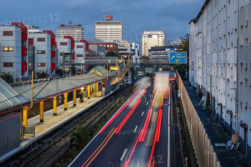 Essen, North Rhine-Westphalia, Germany, motorway A40 in the city centre, exit Essen Zentrum, during dusk. Man waiting at Savignystrasse underground station. On the right, residential buildings behind a noise barrier. There is a LANUV air monitoring station, Essen Kruppstraße 117 (EKRU2), on the right under the lantern. Residents are affected by noise and exhaust fumes. Building with Postbank, Eon and Evonik lettering at the back, Europe