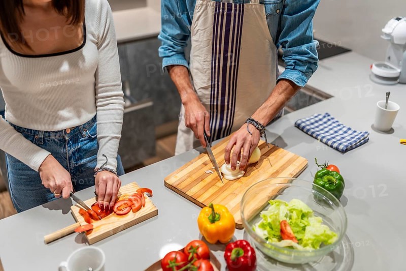 Young couple slicing fresh vegetables on wooden cutting boards, preparing a healthy salad together in a modern kitchen