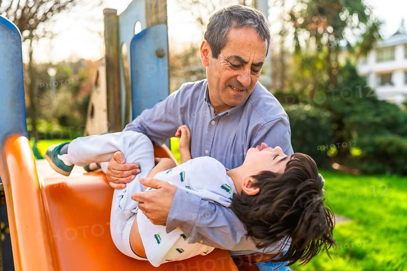 Grandfather catching his grandson sliding down the slide at a playground, having fun together on a sunny day