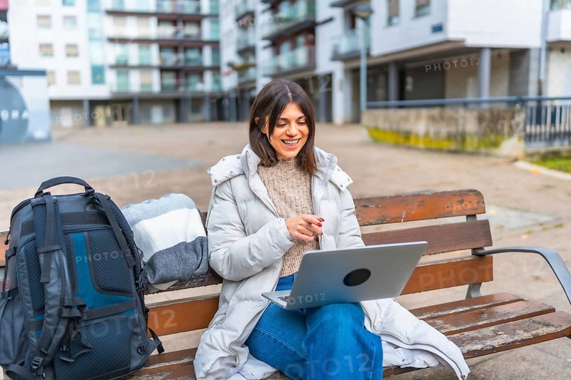 Caucasian smiling woman using laptop sitting on a park in a city square during a cold day