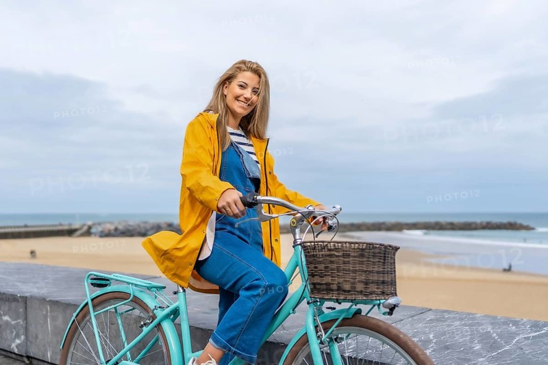 Smiling woman with yellow raincoat sitting on a light blue bicycle by the sea enjoying freedom and healthy lifestyle