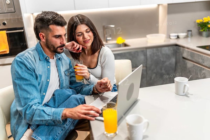 Young couple sitting at kitchen table, drinking orange juice and using laptop, enjoying their time together