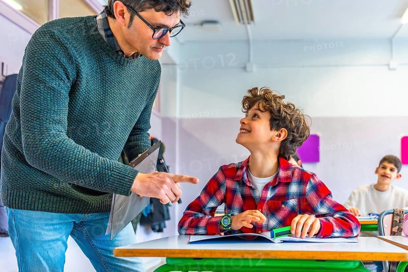 Elementary school teacher engaging with a smiling student while explaining a lesson at their desk in a cheerful classroom setting