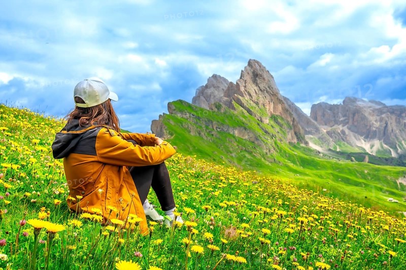 Tourist admiring seceda peaks in the italian dolomites, surrounded by yellow flowers in a sunny summer day