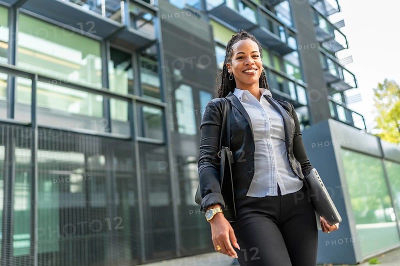 Confident latin modern businesswoman next to block office smiling at camera and carrying laptop and handbag
