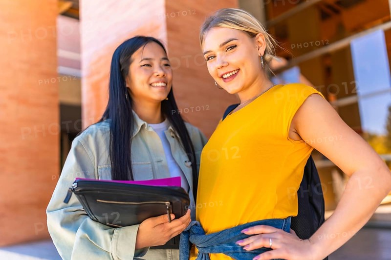 Caucasian and chinese female university students smiling at camera and standing outside the university entrance